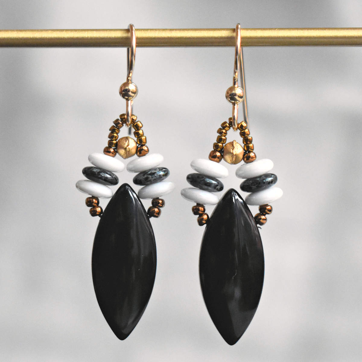 Black and white earrings hanging from a gold bar against a gray background. The earrings are made from black, pointed oval beads suspended from two stacks of black and white discs.