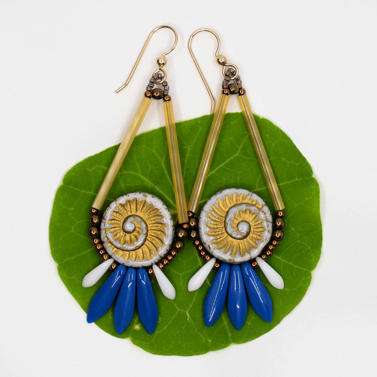 A pair of gold and blue earrings with white spiral shell beads in their centers lay on a circular green leaf and white background. The earrings have gold wires and are formed by two long gold beads on the upper sides, holding white shell impression beads that have gold embossing. At the bottom is a fan of three medium blue dagger beads sandwiched between smaller white daggers.