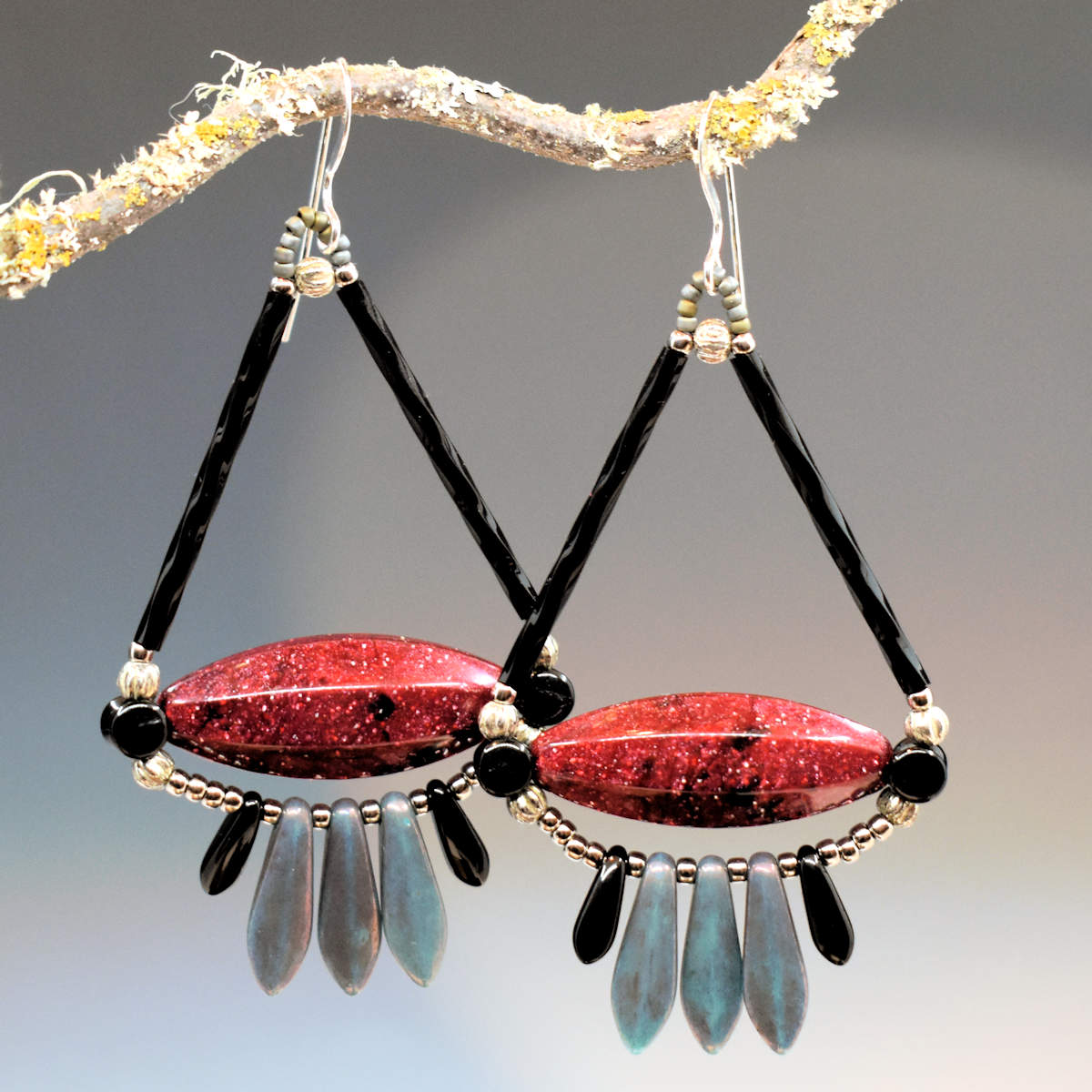 Wide triangular earrings with long black tubes at the top and a base of long, speckled, pointed oval beads hang from a twig. The earrings have silver wires, and below the bottom of the triangle shaped part of the earring is a fringe of five dagger beads, three mottled gray-blue in the center and smaller black beads on either side. 