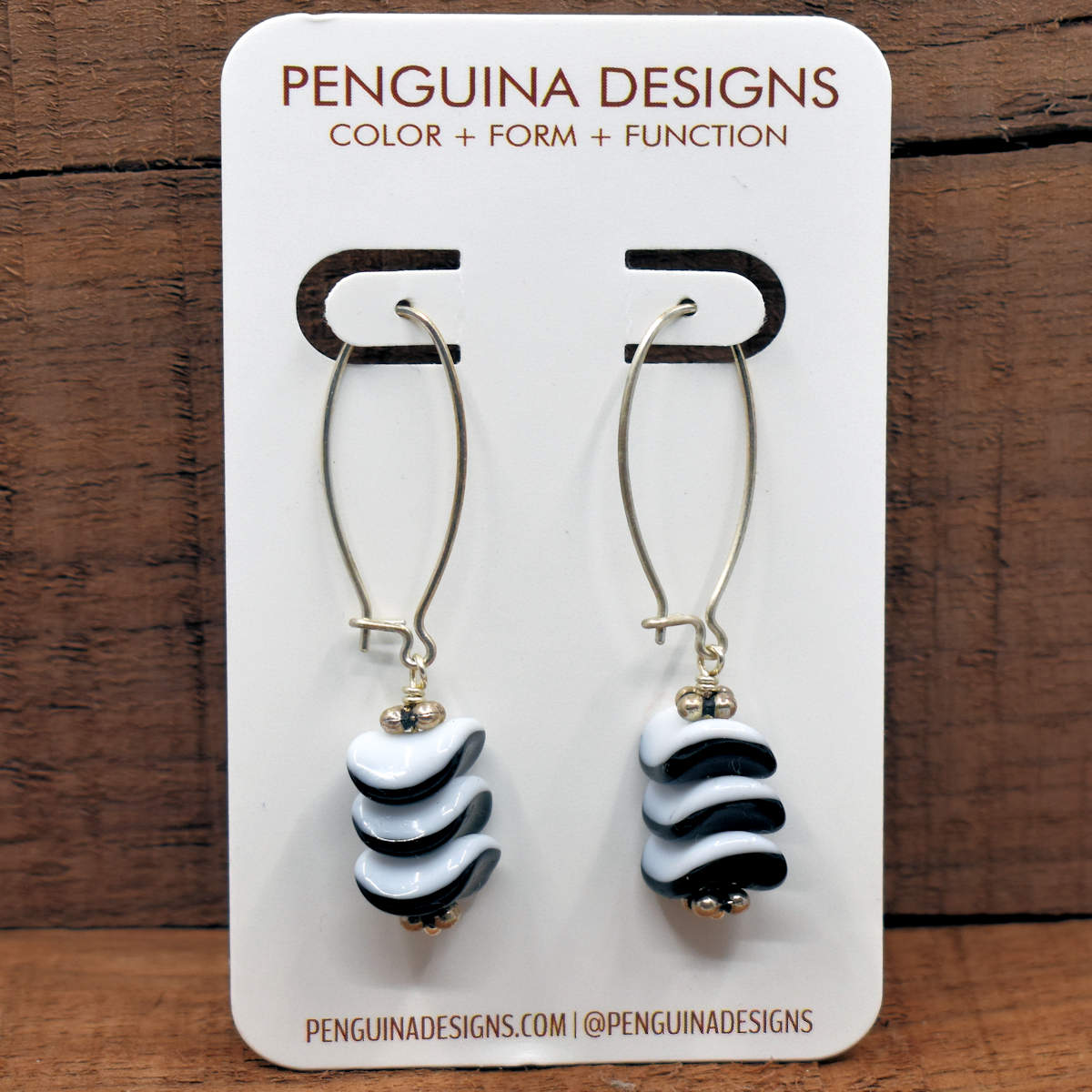 A pair of earrings on a white card rest against a wood background. The earrings have long silver oval wires that latch and the drop part of the earring is a stack of nested wave shaped beads which have black glass on their bottom halves and white on the top.