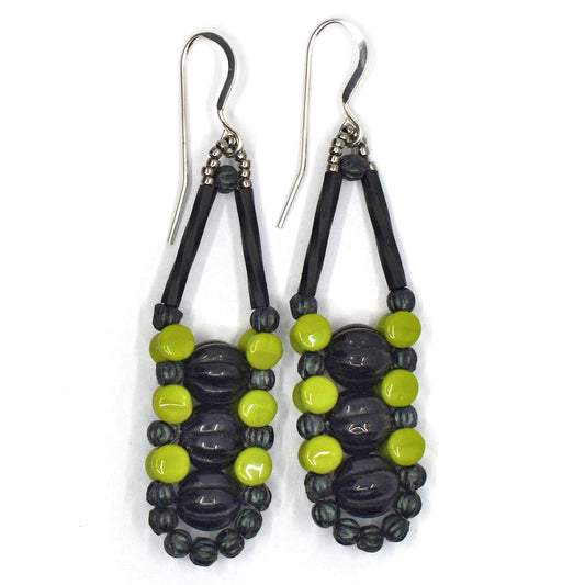 Long earrings with black centers and an outline of dark and light green with silver accents and ear wires on a white background. There are three black beads stacked vertically in the center of these beads, like peas, surrounded by an outline of dark metallic green and avocado green beads.