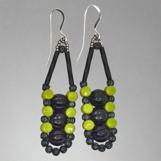 Long earrings with black centers and an outline of dark and light green with silver accents and ear wires on a white background. There are three black beads stacked vertically in the center of these beads, like peas, surrounded by an outline of dark metallic green and avocado green beads.