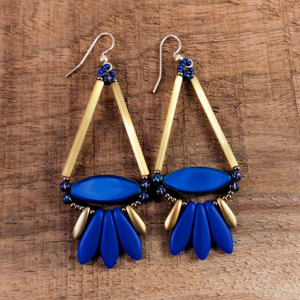 Triangle shaped earrings with a blue and gold fringe lay on a wood background. The earrings have gold ear wires. The triangle shape of the earrings is formed by gold tubes on the upper two sides and a flat blue pointed oval bead at the base. The fringe below the triangle shape is formed with three matte blue daggers sandwiched between smaller gold daggers. 