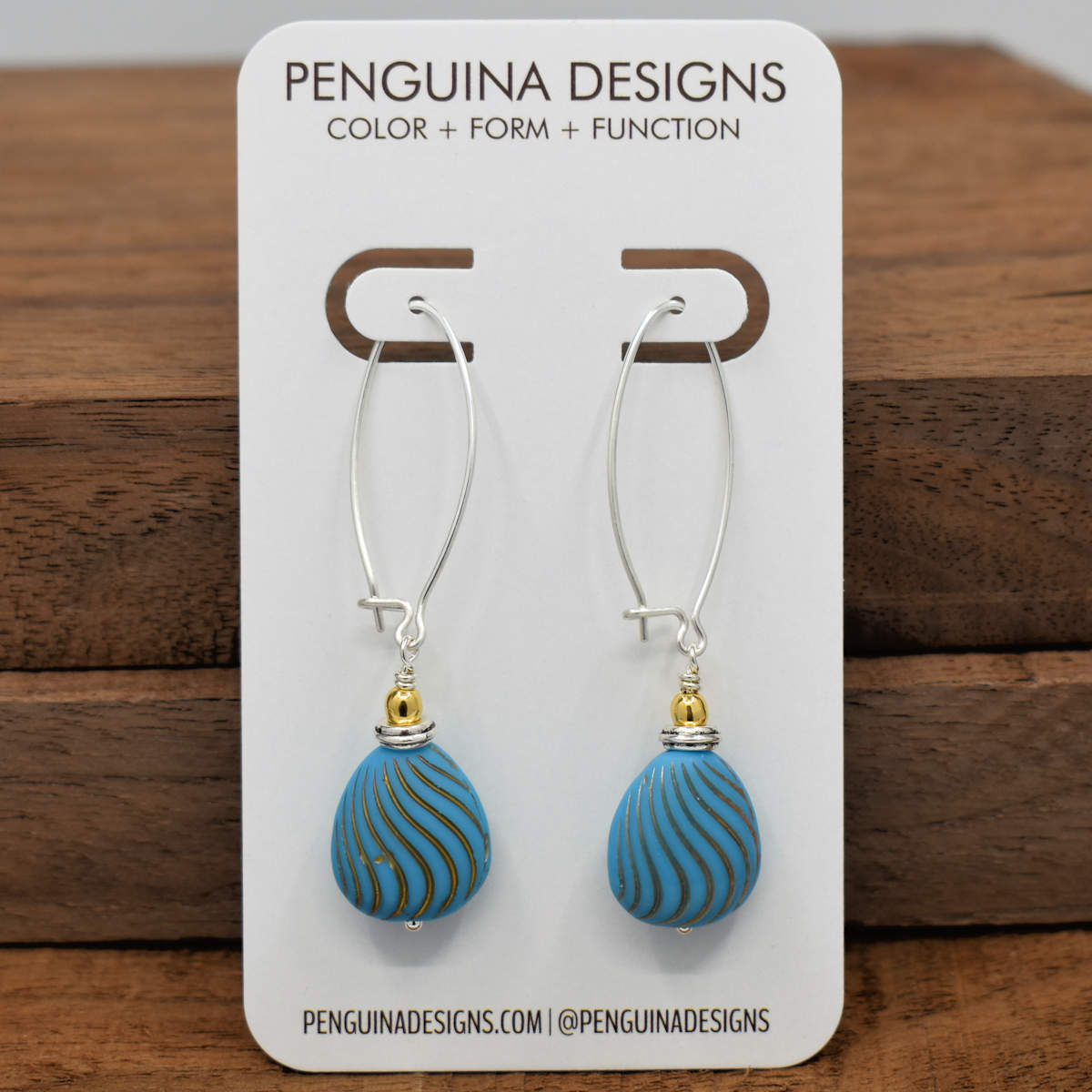 A pair of earrings on a white card rest against a wood background. The earrings have long silver oval wires that latch and a drop at the bottom that is formed from medium blue flat drop beads embossed with wavy gold stripes.