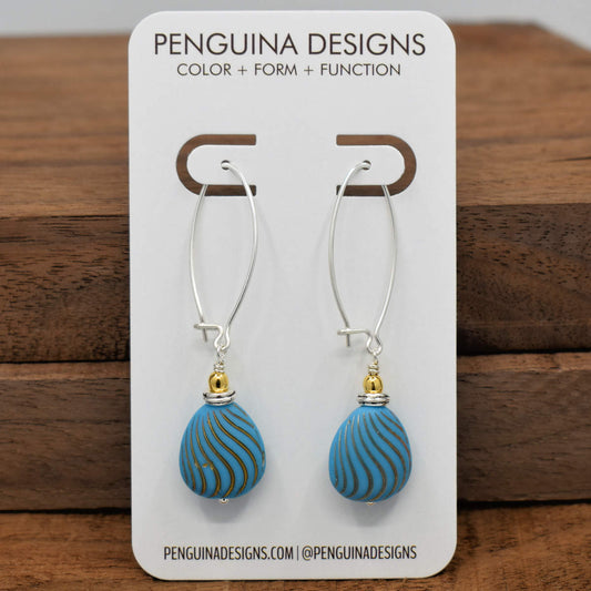 A pair of earrings on a white card rest against a wood background. The earrings have long silver oval wires that latch and a drop at the bottom that is formed from medium blue flat drop beads embossed with wavy gold stripes.