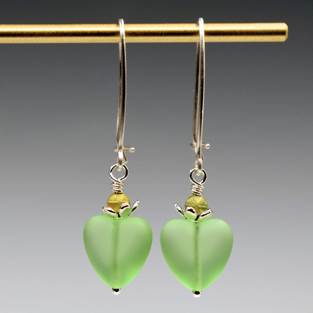 A pair of earrings hang from a gold bar, against a gray background. The earrings have long silver oval wires that latch and small frosted pastel green glass hearts at the bottom.