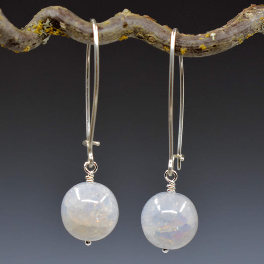 A pair of earrings hang from a twisted twig with lichen on it, against a gray background. The earrings have long silver oval wires that latch and translucent milky white crackle glass balls at the bottom.