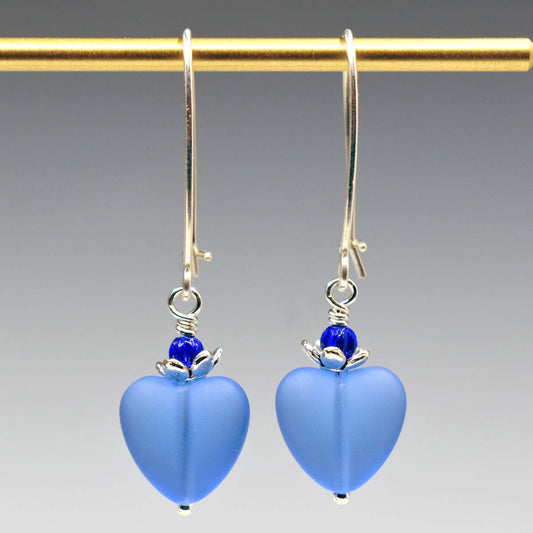A pair of earrings hang from a gold bar, against a gray background. The earrings have long silver oval wires that latch and small frosted translucent pastel blue glass hearts at the bottom.