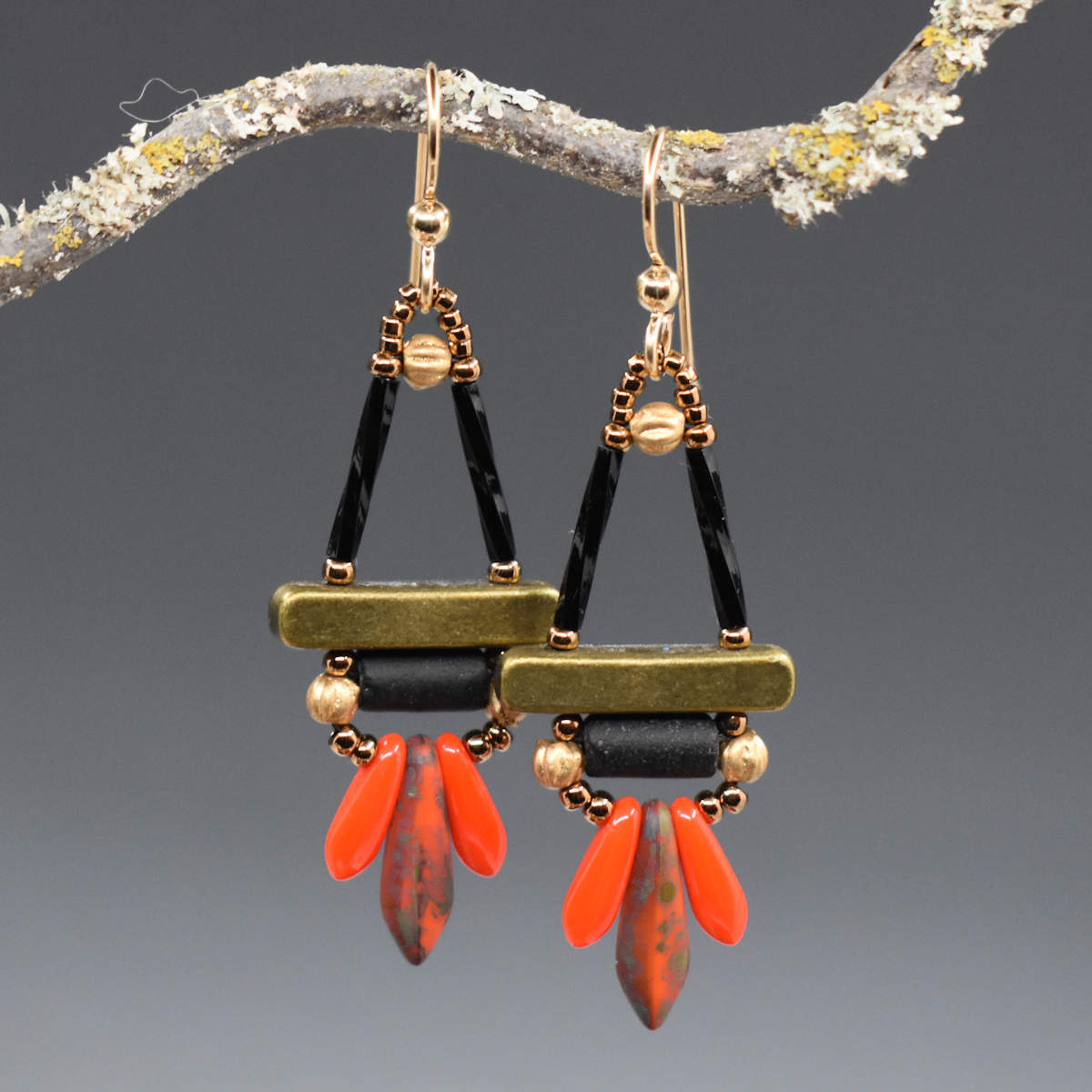 Black, red and gold earrings hang from a lichen covered twig in front of a gray background. The earrings are a triangle shape on top, with two black upright sides and a gold colored bar at the bottom. Below the gold bar is a black tube with gold beads on either side. At the bottom is a spray of three dagger shaped beads, light red smaller ones on the outside and a larger black-speckled red on the inside.