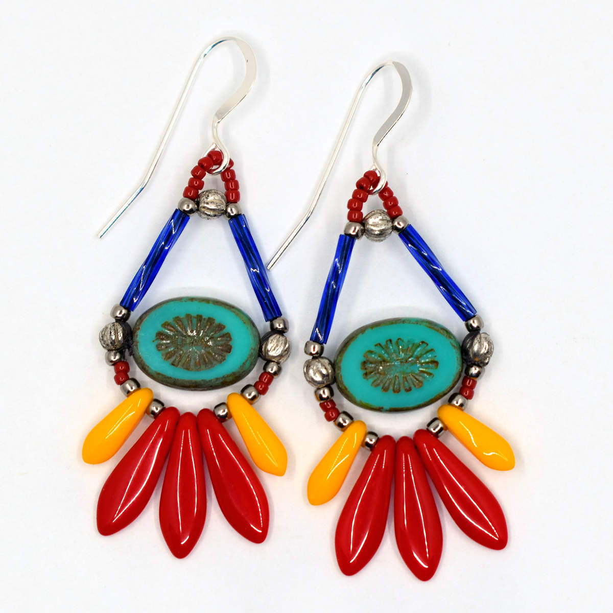 Bright teardrop shaped earrings with silver earwires lay on a white background. The earrings' teardrop shape is formed by blue tube beads at the top and turquoise oval beads with an embossed starburst in the middle. At the bottom is a long fringe of dagger beads, with three larger red ones in the middle and smaller light orange ones framing them. 