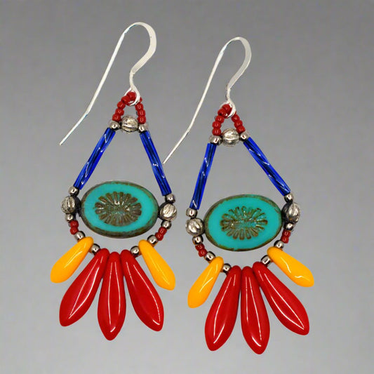 Bright teardrop shaped earrings with silver earwires lay on a white background. The earrings' teardrop shape is formed by blue tube beads at the top and turquoise oval beads with an embossed starburst in the middle. At the bottom is a long fringe of dagger beads, with three larger red ones in the middle and smaller light orange ones framing them. 