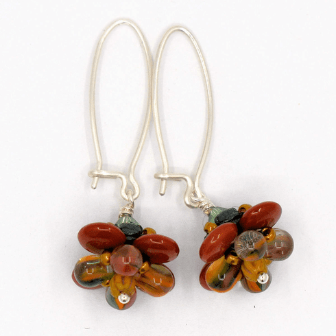 A pair of silver earrings with a brown, mustard and green flower on the bottom lay on a white background. The earrings have long silver oval wires that latch and the dangling parts are beaded flowers with rust colored outer petals and interior petals that are clear with mustard, dark green, and brown stripes.