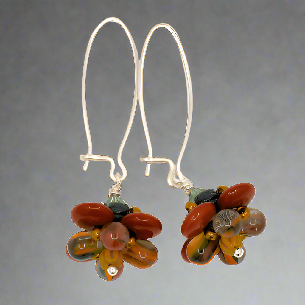 A pair of silver earrings with a brown, mustard and green flower on the bottom lay on a white background. The earrings have long silver oval wires that latch and the dangling parts are beaded flowers with rust colored outer petals and interior petals that are clear with mustard, dark green, and brown stripes.