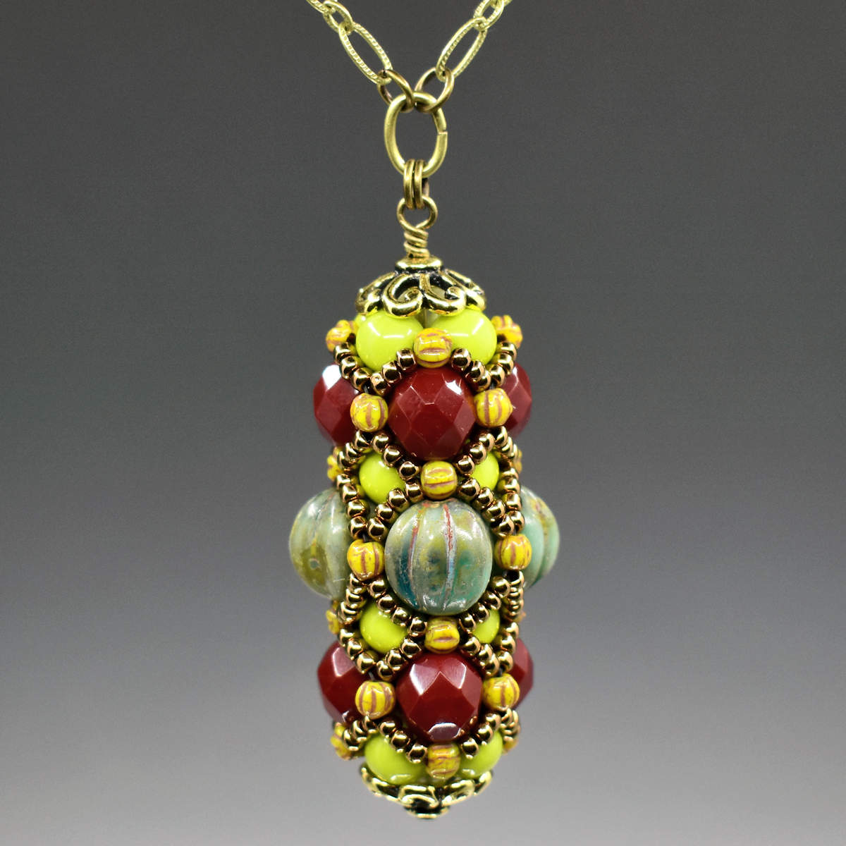 A green and burgundy beaded pendant on a gold colored chain. There is a center row of large mottled turquoise beads for the center row, and burgundy for the outer row.  The chartreuse green beads that form the rest of the base structure show in the spaces between the gold seed bead overlay.