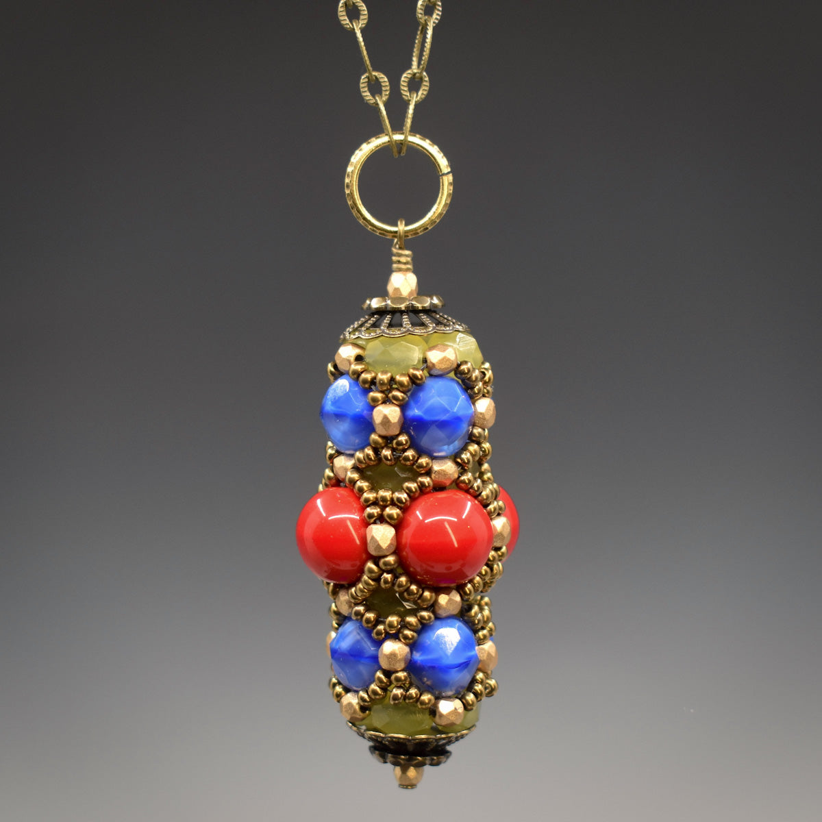 A red, blue, gold and yellow pendant hangs from a gold colored ring. This pendant has a wrm red row of beads in the center and swirly medium blue beads form the outer rows. Pale yellow beads are just visible underneath an overlay of gold seed beads. The pendant is secured between gold colored filigree style bead caps. 