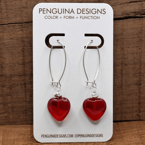 A pair of earrings on a white card rest against a wood background. The earrings have long silver oval wires that latch and  and glossy, rounded hearts.