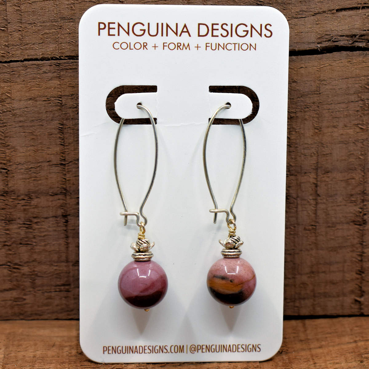 A pair of earrings on a white card rest against a wood background. The earrings have long silver oval wires that latch and dusty pink stone balls at the bottom that have landscape-like layers of darker color at the bottom.