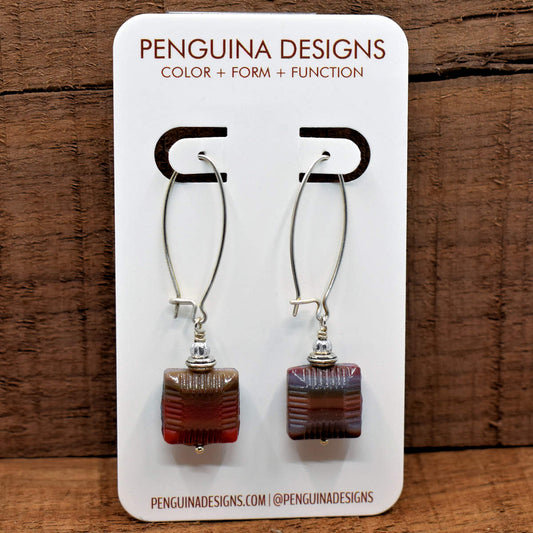 A pair of earrings on a white card rest against a wood background. The earrings have long silver oval wires that latch and square drops at the bottom that have ridged beveled sides. The squares have streaky layers of reds, purples, and gray blue.