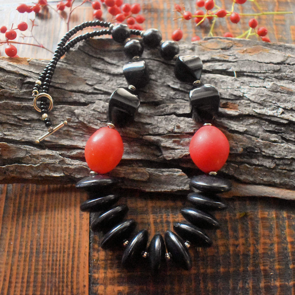 A black and red statement necklace is draped over a wood backdrop with red berries in the background. The necklace has a swath of chunky black discs in the center, bright red ovals, and then a series of black beads that decrease in size, with a few inches of black seed beads at the back. There is a silver toggle clasp.