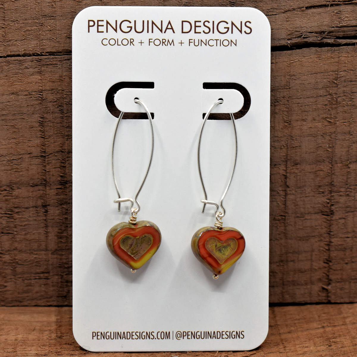 A pair of earrings on a white card rest against a wood background. The earrings have long silver oval wires that latch with heart beads at the bottom that are a vaiegated dark orange and yellow. The hearts are cut to show the colored glass in a wide outline around the outer part, leaving the outside edges and embossed interior heart shape covered in the stone-like tan finish