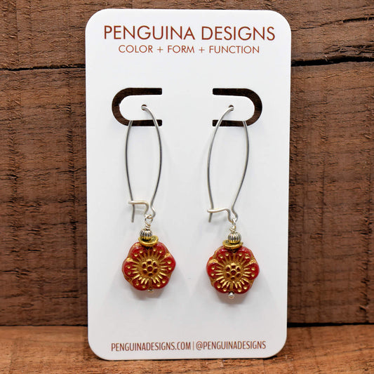 A pair of earrings on a white card rest against a wood background. The earrings have long silver oval wires that latch and red six petaled flowers at the bottom with embossed gold accents.