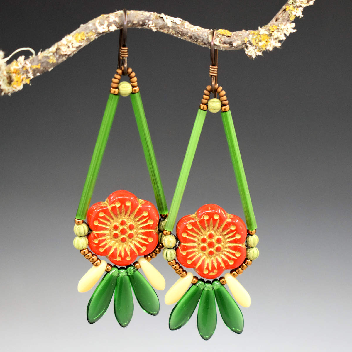 Long green earrings with a red flower center hang from a lichen covered twig. The earrings are shaped like a teardrop with tail feathers at the bottom. The teardrop shape is formed by two long green tube beads and the round part is a warm orange flower with gold embossed details. At the bottom are three transparent green dagger beads with a smaller cream dagger on either side. The earrings have dark ear wires and gold seed bead accents.