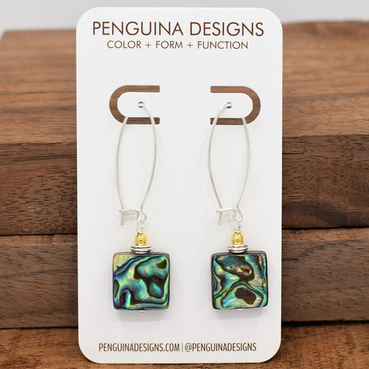 A pair of earrings on a white card rest against a wood background. The earrings have long silver oval wires that latch and iridescent blue green shell squares at the bottom.