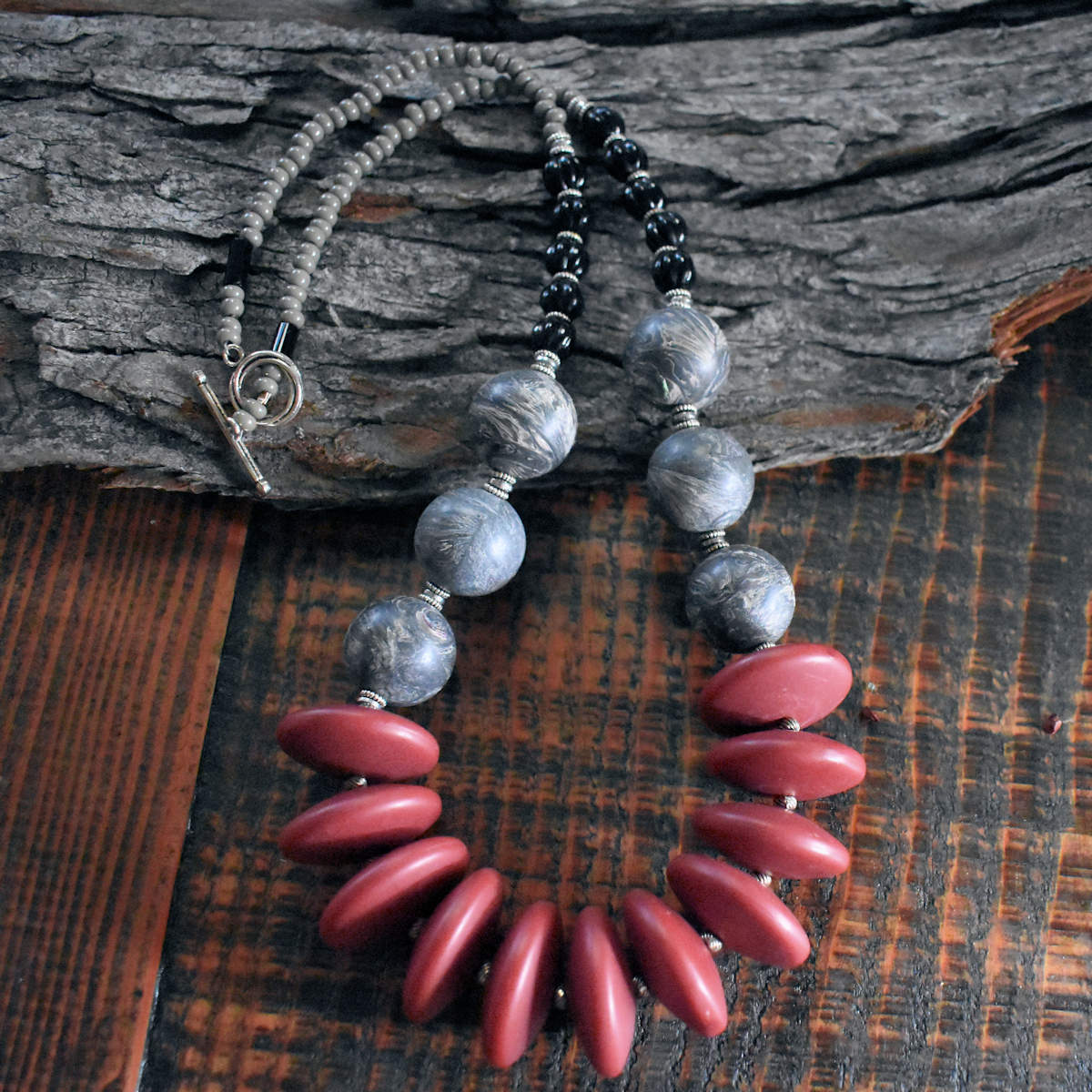 A chunky necklace with mauve and gray beads is draped on a wood backdrop. The necklace has eleven large mauve disc beads in the center, then three gray and cream swirled beads on each side. The necklace is finished with five larger black beads and a few inches of gray seed beads. The clasp is a silver toggle.