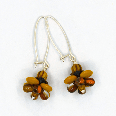 A pair of silver earrings with mustard and brown flowers on a white background. The earrings have long silver oval wires that latch and  dangling beaded flowers with mustard outer petals and inner petals made of clear glass with orange and brown stripes inside of it..