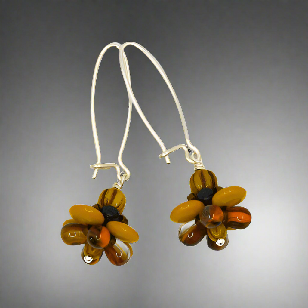 A pair of silver earrings with mustard and brown flowers on a white background. The earrings have long silver oval wires that latch and  dangling beaded flowers with mustard outer petals and inner petals made of clear glass with orange and brown stripes inside of it..