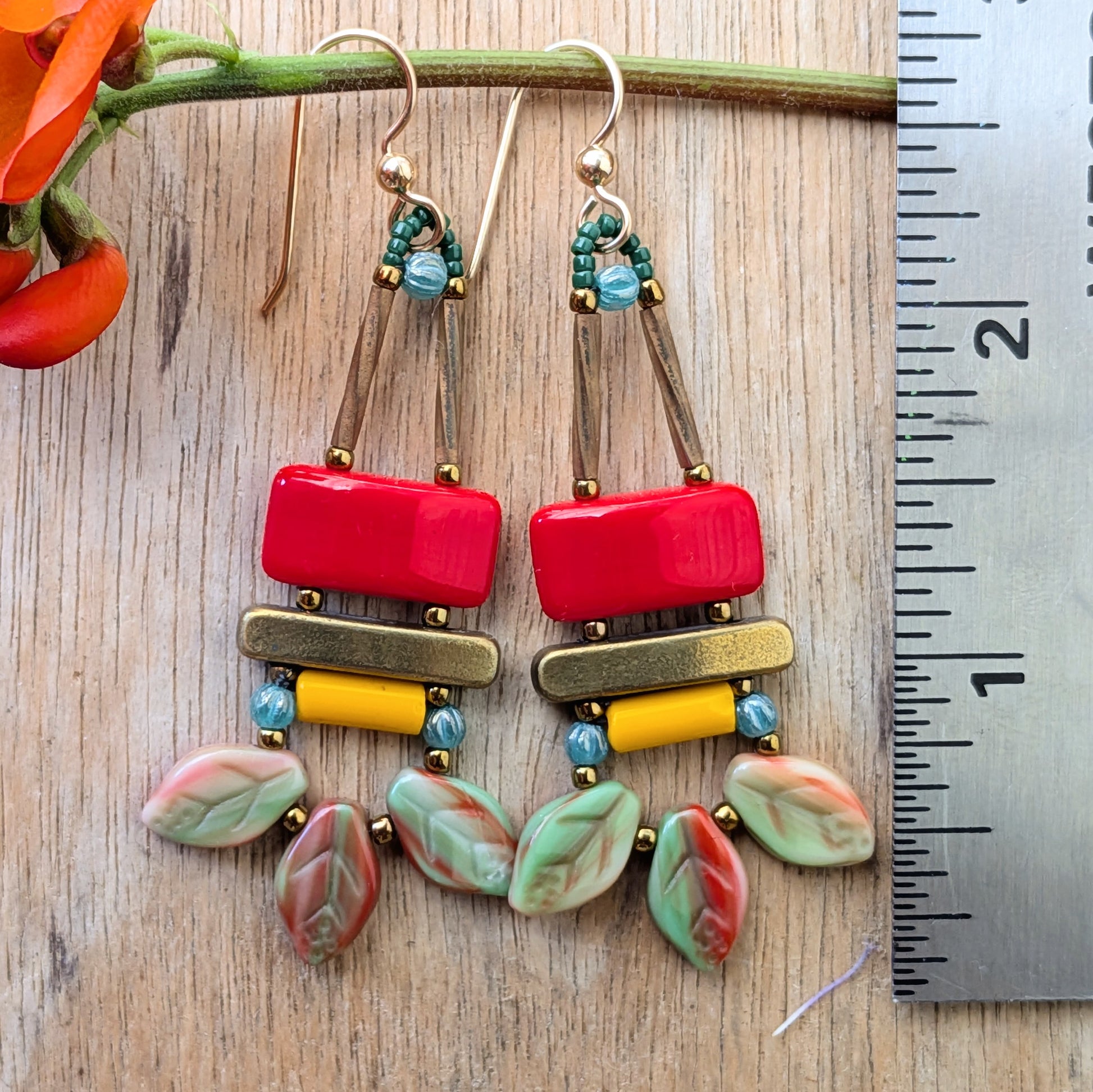 Colorful earrings on a wood background with a stem and flowers and a rule for scale. The earrings  have a red rectangle stacked on top of a brass colored bar, with a light orange tube and swag of three red and green streaked leaves at the bottom.