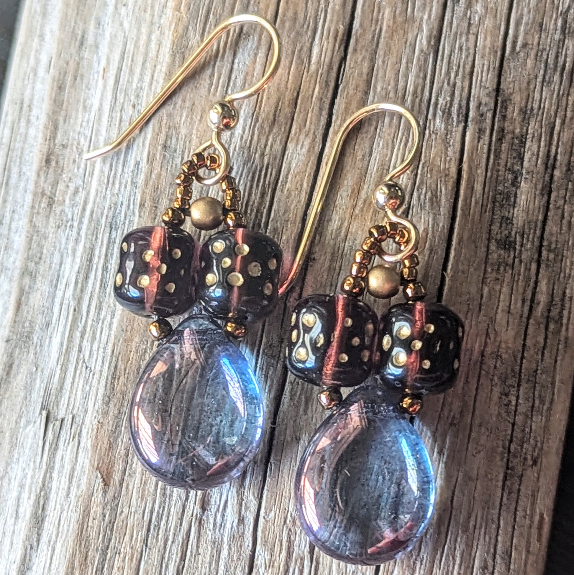Pair of earrings with purple teardrop beads, plum cylinder beads, and gold accents on a wooden surface.
