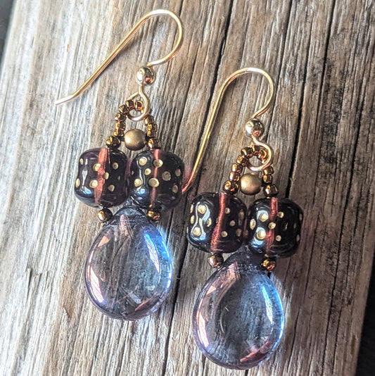 Pair of earrings with purple teardrop beads, plum cylinder beads, and gold accents on a wooden surface.