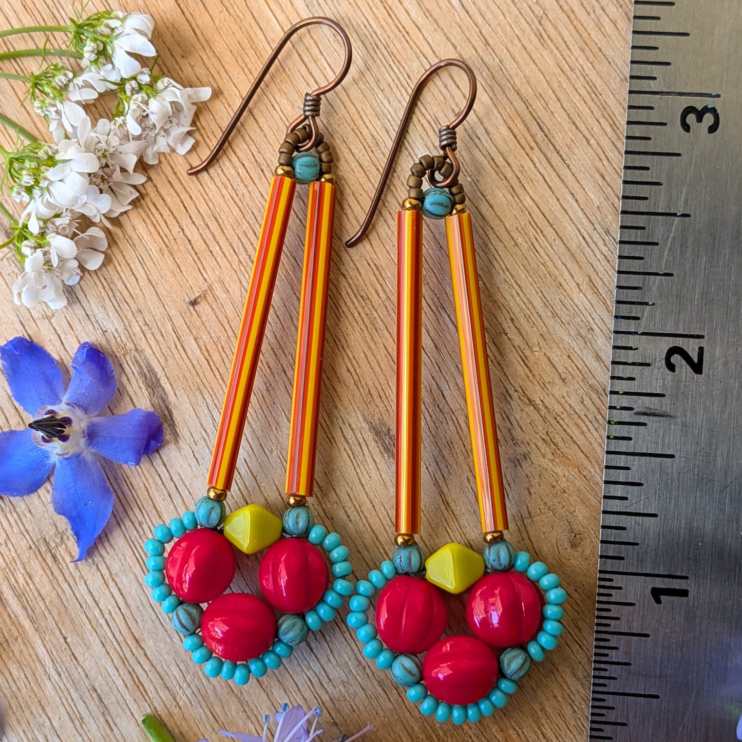 Brightly colored earrings on a wood background with flowers and a ruler for scale. The earrings have a clover of red beads outlined in tiny turquoise beads. The clover shape is suspended from a pair of long, thin red and orange striped tube beads.