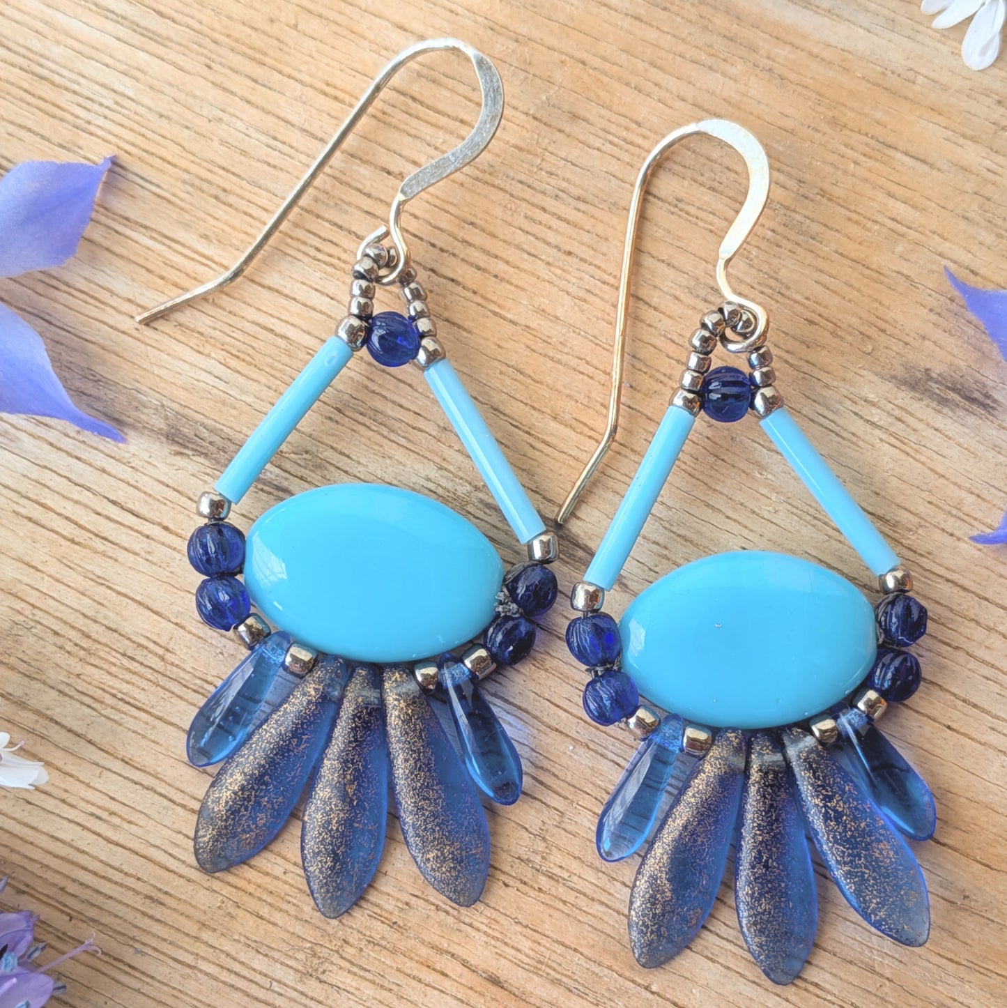 Blue and silver earrings on a wooden surface with flowers. These earrings have a light blue oval suspended by a triangle of light blue bugle beads at the top and a swag of shimmery darker blue beads around the bottom.