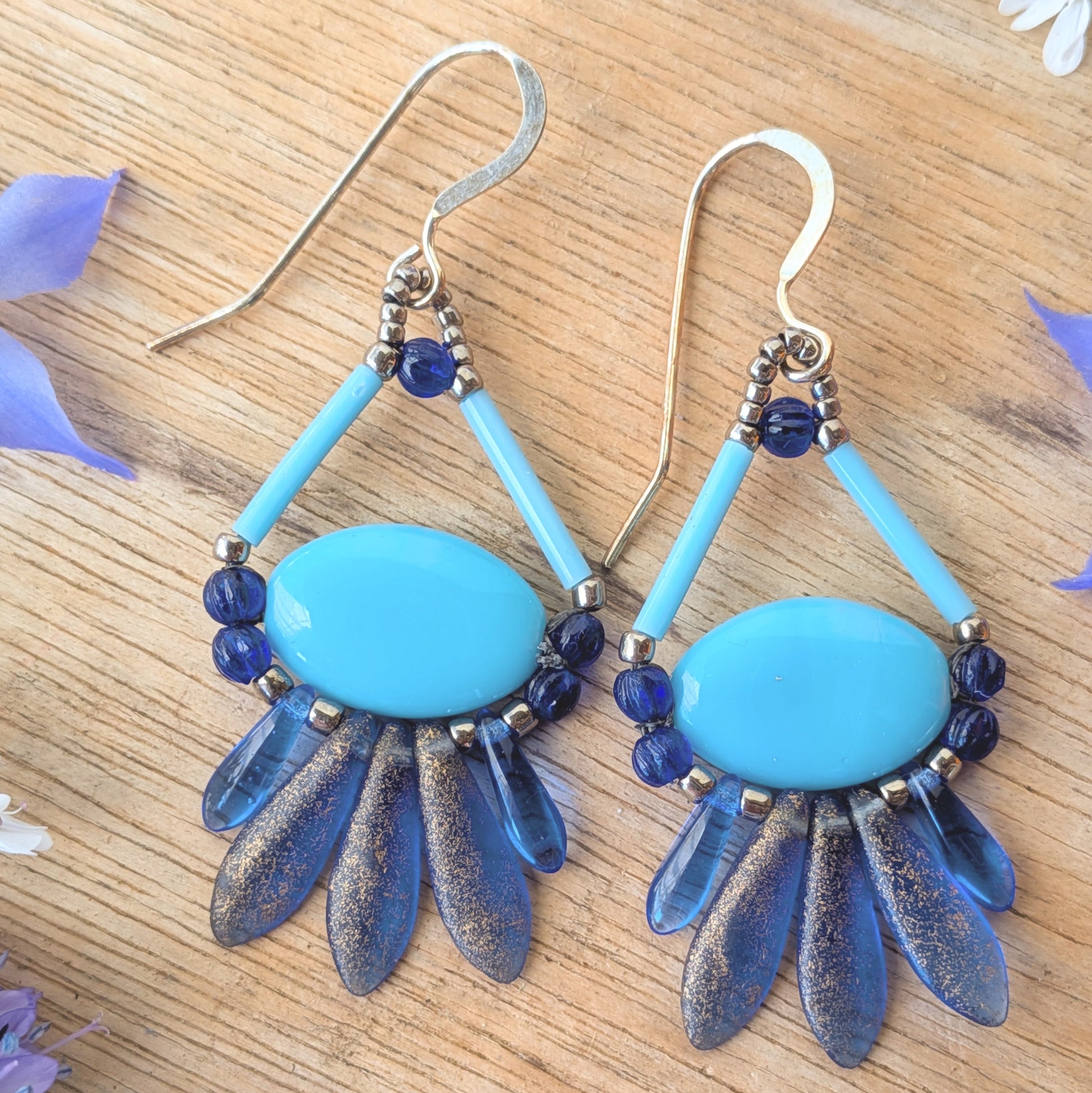 Blue and silver earrings on a wooden surface with flowers. These earrings have a light blue oval suspended by a triangle of light blue bugle beads at the top and a swag of shimmery darker blue beads around the bottom.