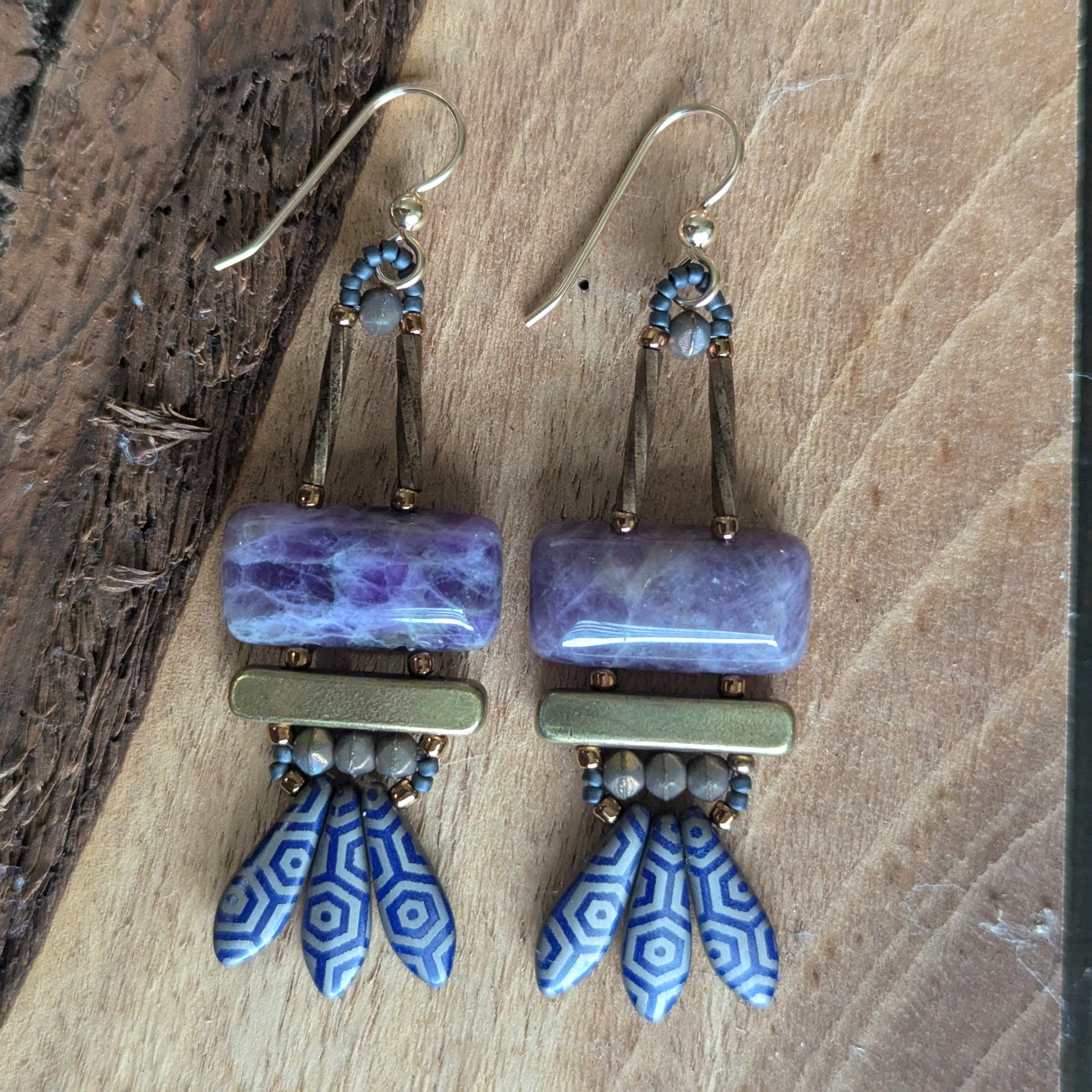 Purple earrings on a slice of wood. The earrings have purple stone rectangles on top with gold bars and navy blue daggers with a silver-gray octagon pattern.