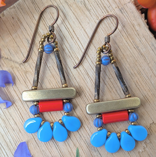 Blue and red earrings with petal shaped beads around the bottom, on a wood background with flowers. 