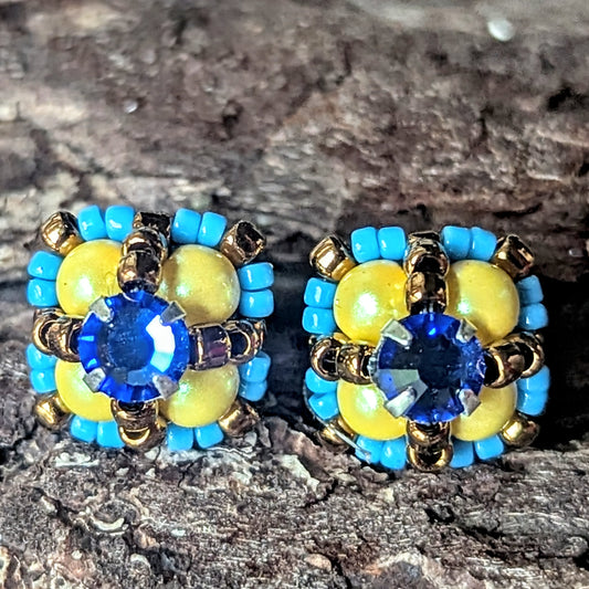 Four-lobed stud earrings with yellow beads surrounded by smaller turquoise beads and a blue rhinestone center, on a wood bark background.