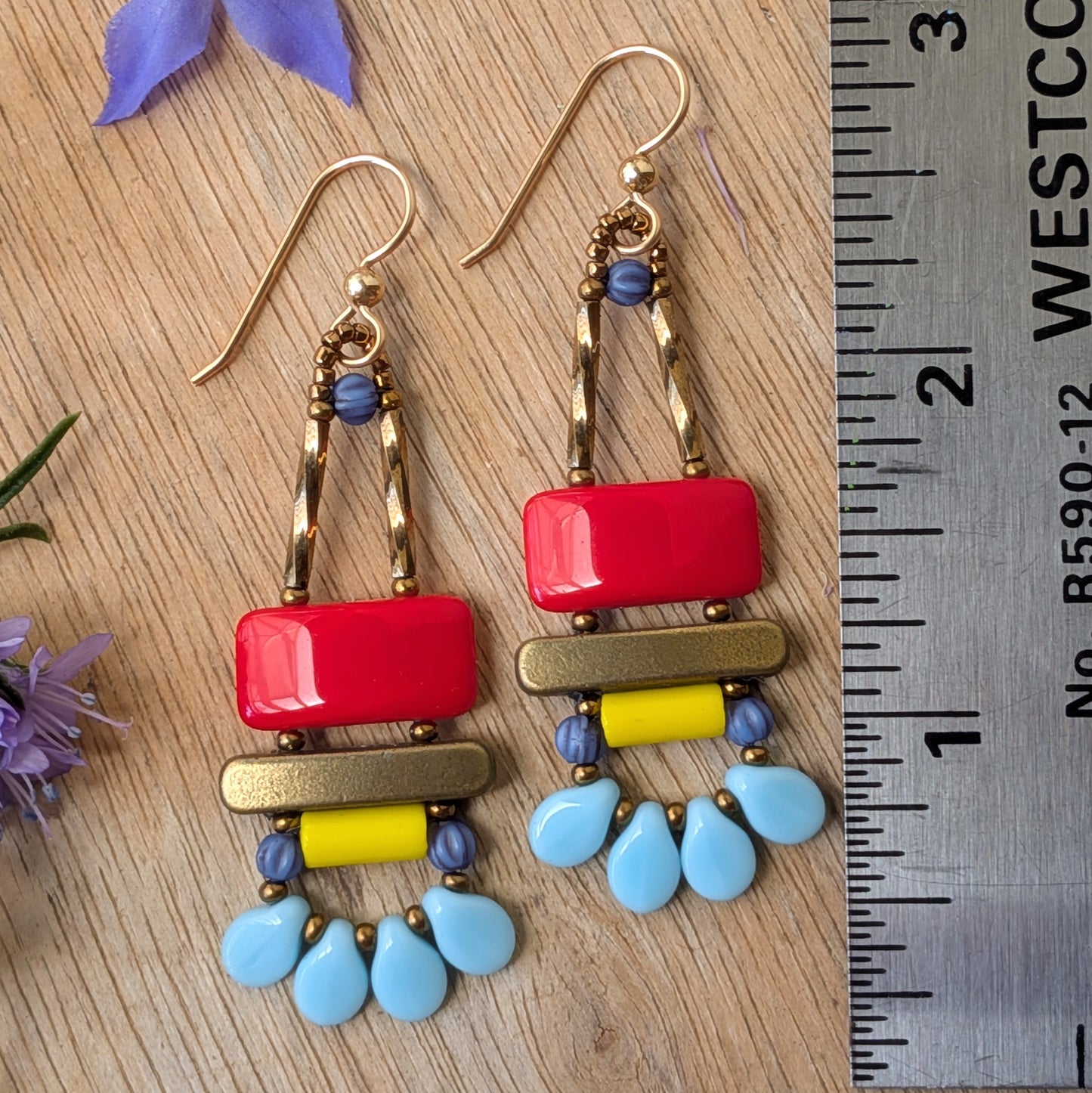 Colorful earrings on a wood background next to a ruler. The earrings are a red rectangle on top of a gold bar and below the bar, a yellow tube with a swag of four light blue petals.