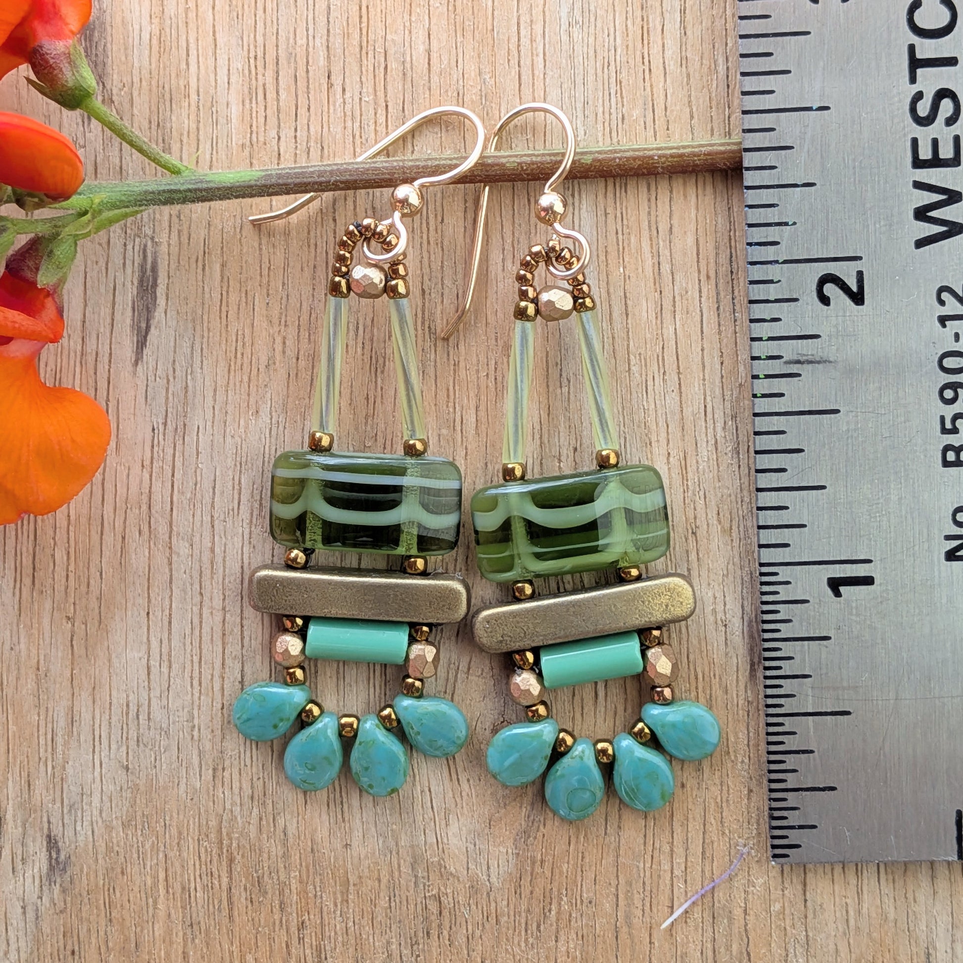 Green earrings on a wood background with a ruler for scale. Earrings with a white-striped, green rectangle on top of a gold bar, mint tube bar and a swag of four mottled turquoise petals.