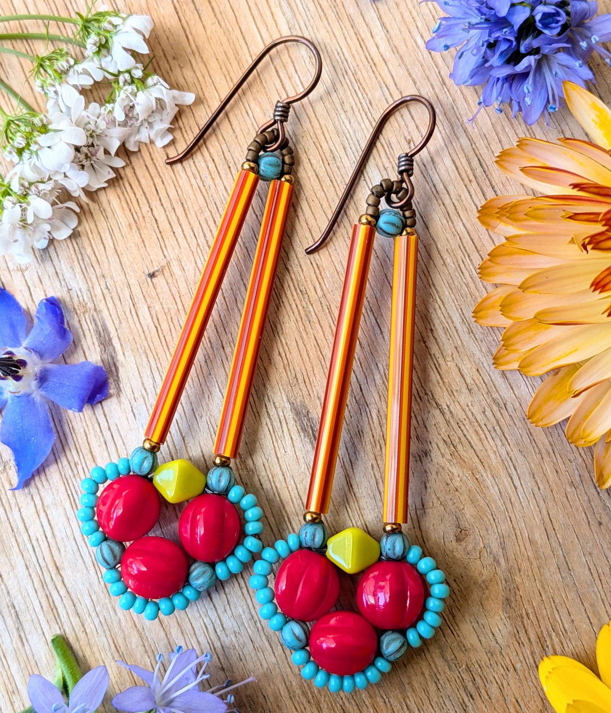 Brightly colored earrings on a wood background with flowers. The earrings have a clover of red beads outlined in tiny turquoise beads. The clover shape is suspended from a pair of long, thin red and orange striped tube beads.