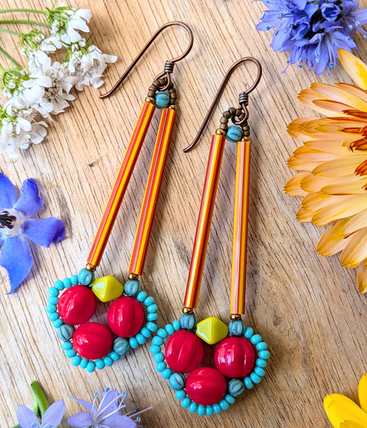 Brightly colored earrings on a wood background with flowers. The earrings have a clover of red beads outlined in tiny turquoise beads. The clover shape is suspended from a pair of long, thin red and orange striped tube beads.