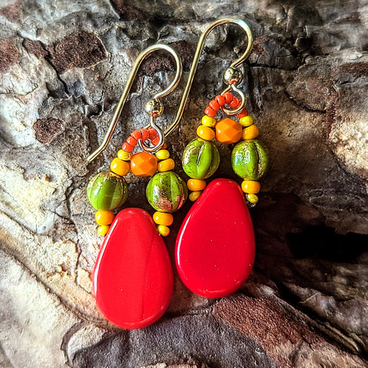 Colorful beaded earrings with red teardrop-shaped beads accented with green, yellow and orange beads on a wood bark background.