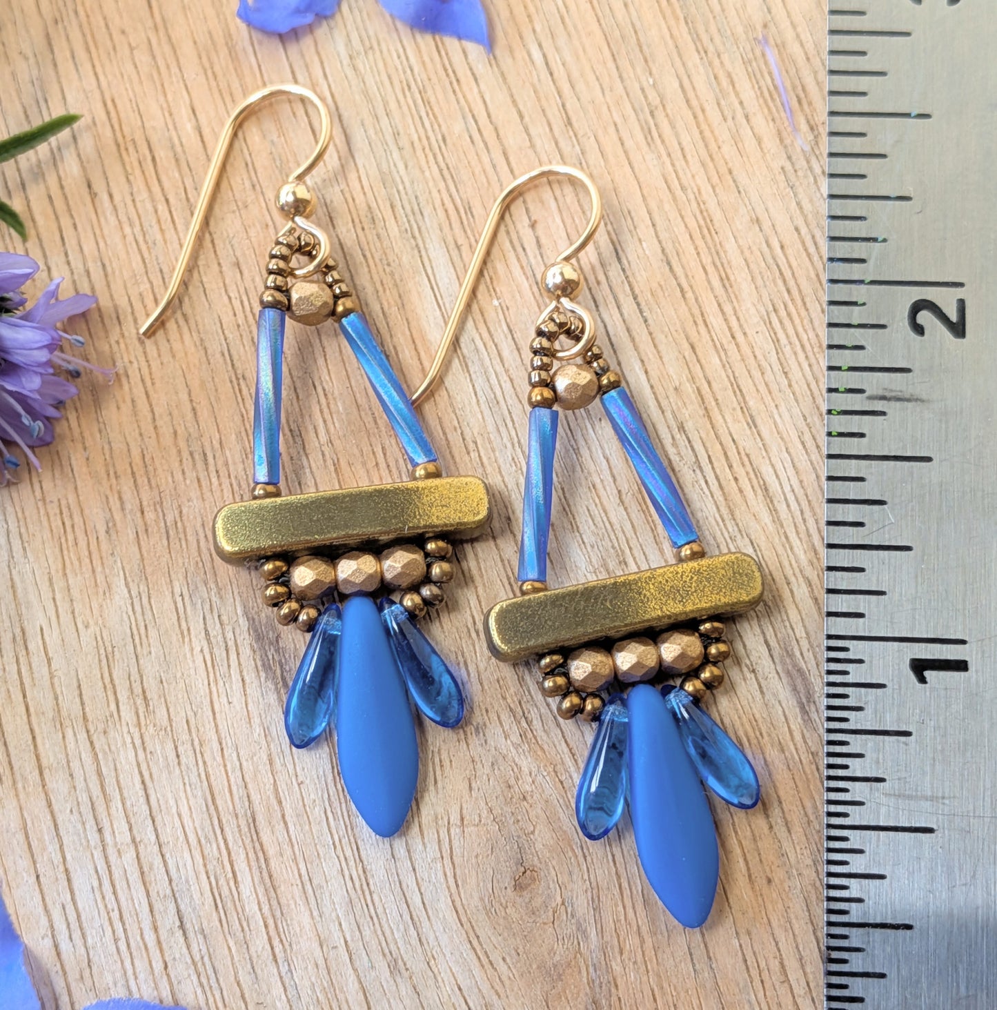 Blue and gold earrings with long blue dagger beads at the bottom, on a wood background, with flowers and a ruler for scale.