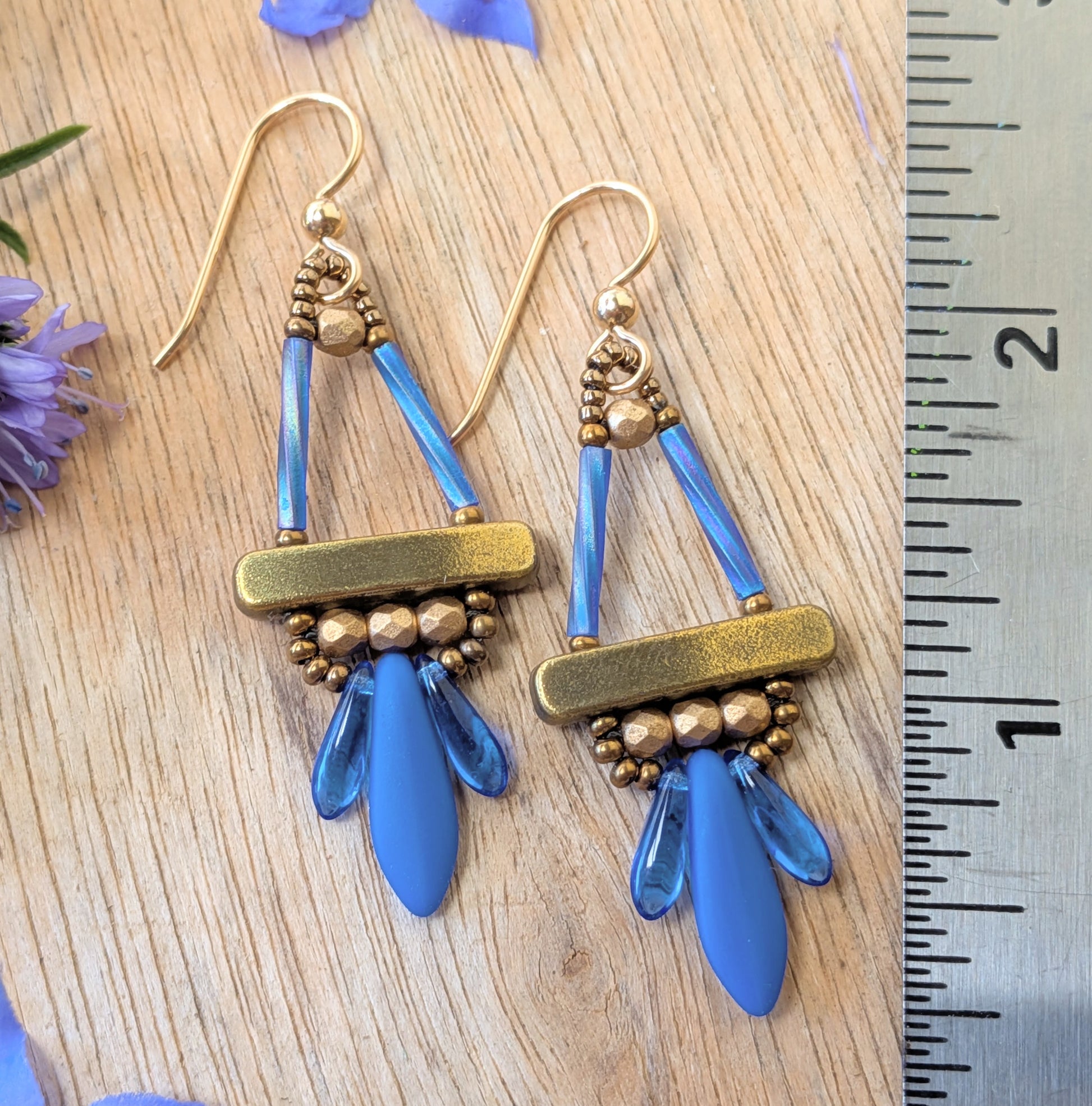 Blue and gold earrings with long blue dagger beads at the bottom, on a wood background, with flowers and a ruler for scale.