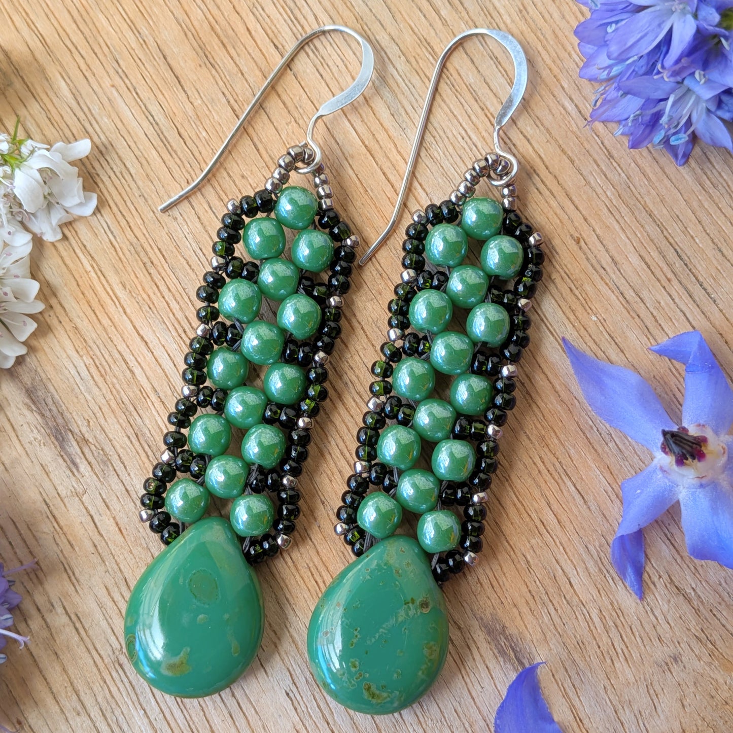 Pine green beaded earrings with teardrops at the bottom and a laced outline of darker green seed beads, resting on a wood background.