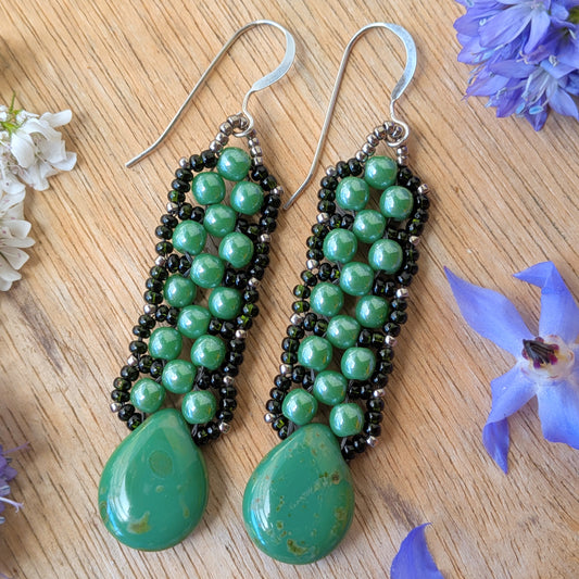 Pine green beaded earrings with teardrops at the bottom and a laced outline of darker green seed beads, resting on a wood background.