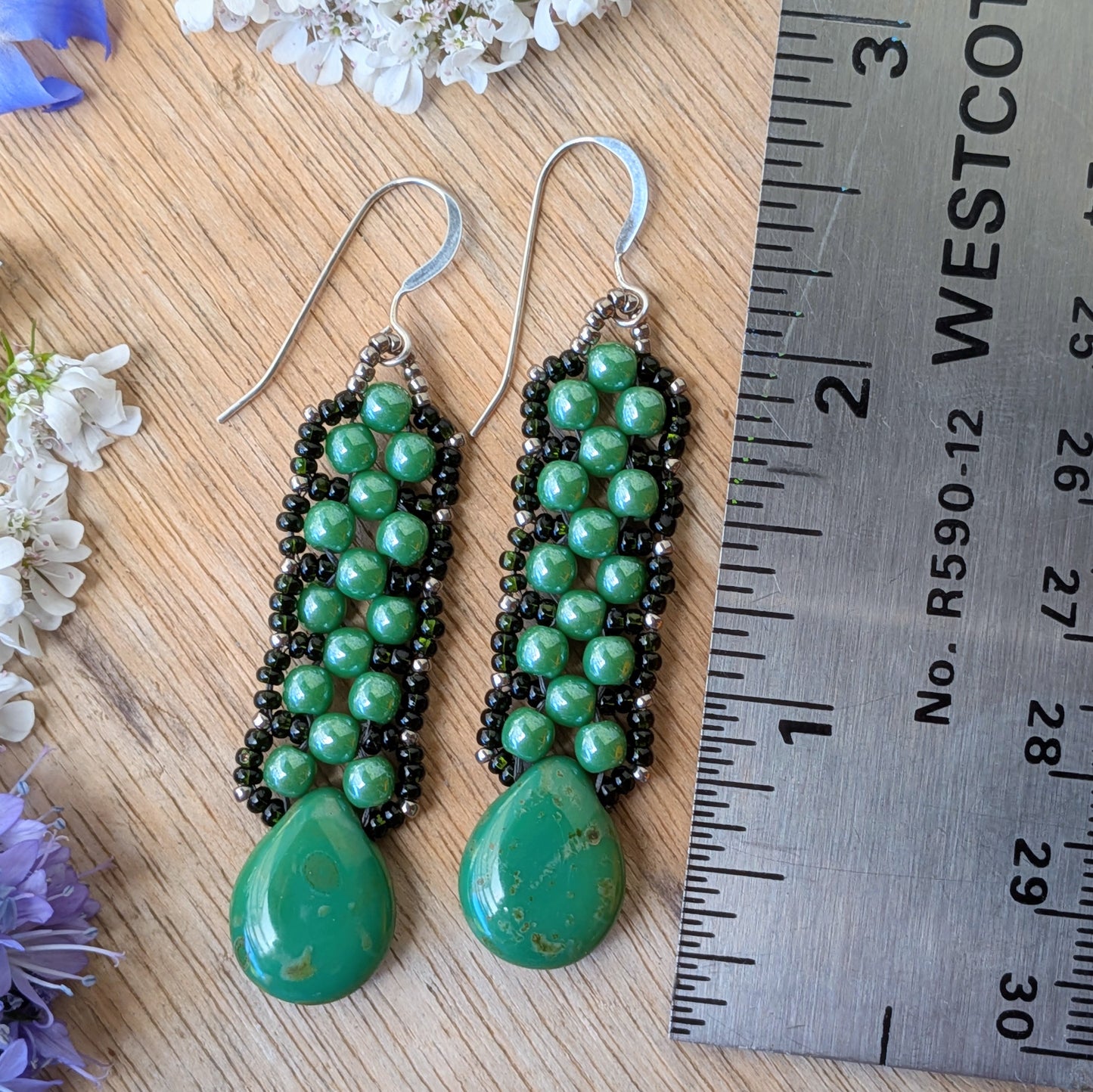 Pine green beaded earrings with teardrops at the bottom and a laced outline of darker green seed beads, resting on a wood background, with a ruler for scale.