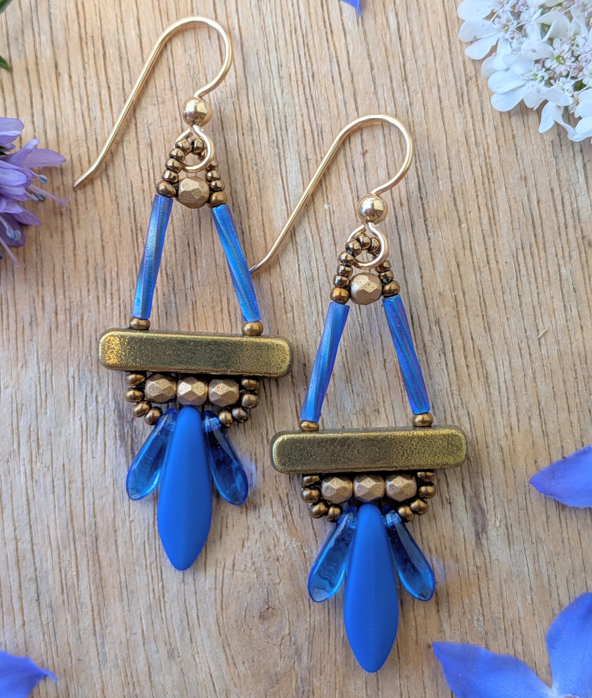 Blue and gold earrings with long blue dagger beads at the bottom, on a wood background, with flowers.